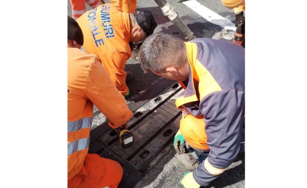 Construction workers in high-visibility orange gear operating a handheld device over an open road trench area.