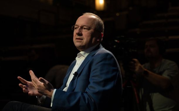 Man in a blue suit being interviewed, gesturing with his hands while speaking, in a dim studio with a camera operator in the background.