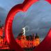 Acrobat on a hoop suspended by cables inside a large red inflatable ring, with a warmly lit historic building in the background at dusk.