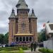 Brick church with green-tiled towers and a yellow awning; crowd gathered at entrance on a cloudy day.