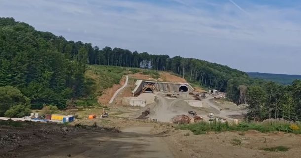 Construction site on a hillside with a tunnel entrance under an embankment, dirt roads and equipment nearby