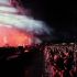 Crowd watches a live concert at night as bright stage lights and smoke illuminate performers on stage ahead.