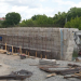 Construction site beside a river with a large reinforced concrete wall, rebar, and an excavator at work as workers move lumber and steel rods.