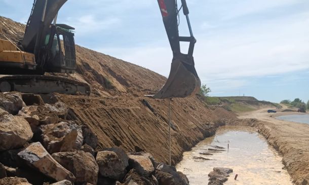 Excavator on a rocky slope digging a long trench along a waterline, with muddy banks and a cloudy sky.