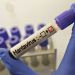 Lab technician in blue gloves holding a test tube labeled 'Hantavirus' with a red checkmark in a laboratory setting.