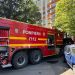 Red fire truck with yellow stripes labeled POMPIERII 112 parked on a city street beside a blue car with a residential building in the background and trees nearby.