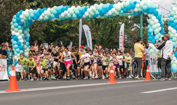 Children start a road race under a blue‑green balloon arch; spectators and organizers watch nearby.