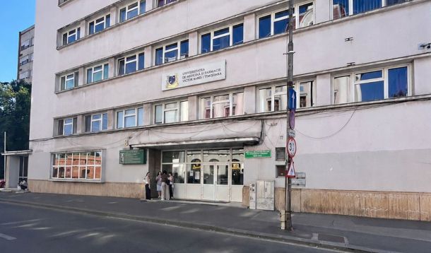 Front view of a white multi-story medical university building with people gathered at the entrance on a city street.