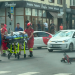 Paramedics in red uniforms wheeling a patient on a stretcher across a city street as a red motorcycle lies on its side nearby, with bystanders and storefronts in the background.