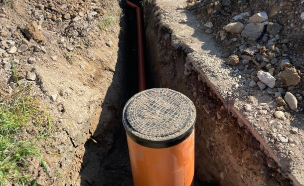 Open trench with an orange PVC pipe and a circular manhole/access lid at a construction site.