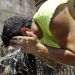 Man in a neon sleeveless shirt splashes water on his head from his hands at an outdoor water fountain, with a coworker in a safety vest nearby.
