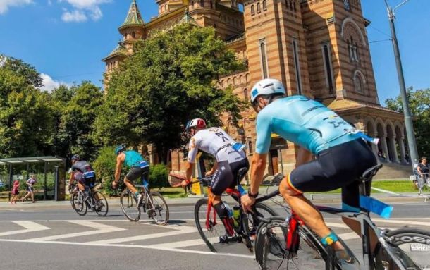 Cyclists ride in a group along a sunny city street past a red-brick historic building with green trees nearby.