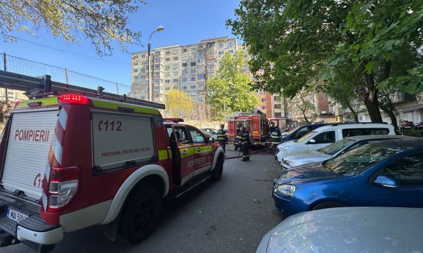 Firefighters in turnout gear work near red fire trucks in a residential parking lot beside apartment buildings.