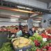 Indoor market stall with fresh vegetables displayed in crates; a vendor wearing headphones sits behind the produce while shoppers browse in the background.