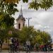 Group of cyclists riding along a tree-lined street with a church steeple visible in the background.
