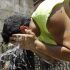 Man in a neon sleeveless shirt splashes water on his head from his hands at an outdoor water fountain, with a coworker in a safety vest nearby.