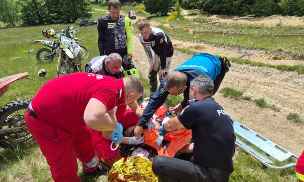 Emergency responders in red and dark uniforms attend to a person on the ground in a grassy off-road area, with dirt bikes nearby.