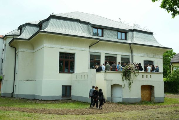 Group of people gathered on a balcony in front of a cream-colored house with a gray tiled roof and decorative molding.