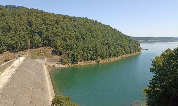 Reservoir scene: a sloped concrete dam on the left, calm turquoise water, and a dense forested hill rising behind.