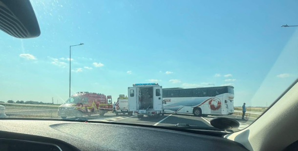 View from inside a car of a highway accident: a white and blue bus on the shoulder, a red emergency vehicle nearby, and a person standing by on a sunny day.