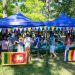 Outdoor park festival under blue canopies with colorful pennant bunting and people browsing vendor tables; Sri Lanka flag visible at front fence.