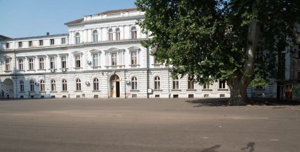 White neoclassical building with arched windows and decorative details, set in an empty plaza with a large tree to the right.