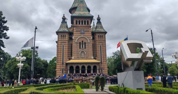 Brick church with green-tiled towers and a yellow awning; crowd gathered at entrance on a cloudy day.