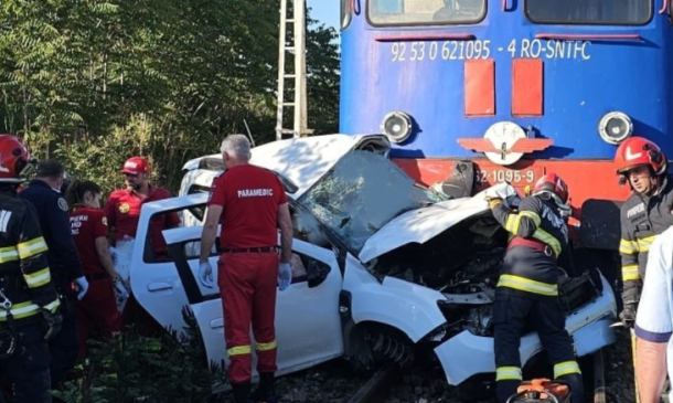 White car severely damaged after collision with a blue train, emergency responders in red and yellow gear assisting on the tracks.