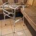 Folding aluminum walker with rubber tips set up in a tiled living room beside a beige sectional sofa.