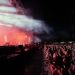 Crowd watches a live concert at night as bright stage lights and smoke illuminate performers on stage ahead.