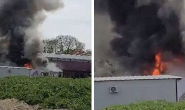 Industrial fire: thick black smoke and flames billow from a low-roof building near a gravel lot and trailers.