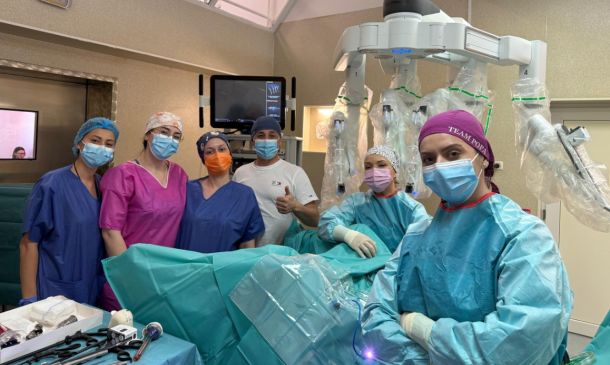 Surgical team in masks and gowns pose in an operating room beside a robotic surgery system with multiple robotic arms.