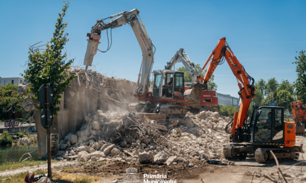 Two excavators demolish a concrete wall at a sunny construction site, dust rising over a pile of rubble; Primăria Municipiului Timișoara logo visible at the bottom.