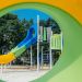 Colorful playground structure with a blue slide and green railings seen through a yellow circular tunnel outdoors, with trees and buildings in the background.