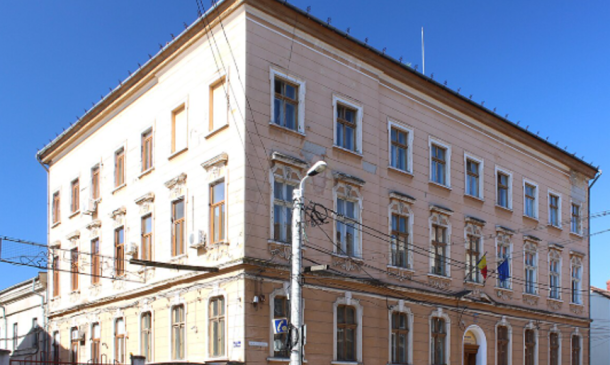 Three-story beige-orange building with decorative window frames and an arched entrance, flags by the facade, under a clear blue sky.