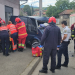 Firefighters and paramedics surround a severely damaged blue van after a street crash, assisting a patient as a crowd stands nearby with a fire truck visible in the background.