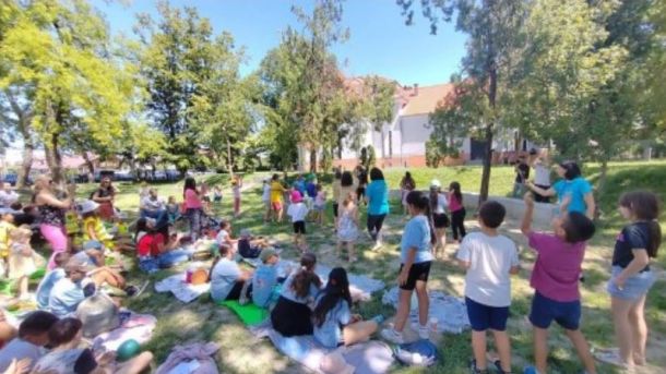 Large group of kids and adults in a sunny park, sitting on blankets and standing, participating in an outdoor activity.