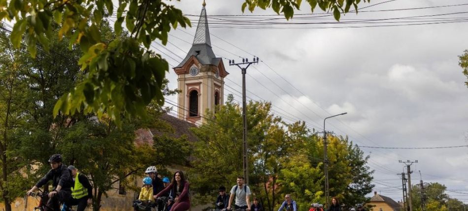 Group of cyclists riding along a tree-lined street with a church steeple visible in the background.