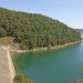 Reservoir scene: a sloped concrete dam on the left, calm turquoise water, and a dense forested hill rising behind.