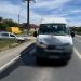 White van in the foreground on a two-lane rural road after an accident; blurred front bumper and a silver car off the left roadside, blue sky above.