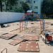 Playground under construction with red and blue looping arches, stacked mats, and construction equipment on a paved area.