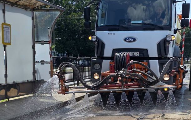 Front view of a white Ford street-cleaning truck spraying water through a bar with multiple nozzles onto the street surface.
