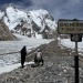 Climbers on a rocky, snow-dusted trail with a Gasherbrum Base Camp sign and towering snowy peaks in the background.
