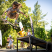 Skater launches off a ramp, mid-air, performing a kickflip while others watch in a sunny park.