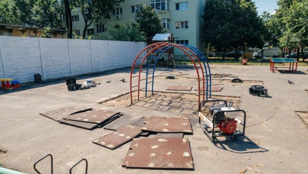 Playground under construction with red and blue looping arches, stacked mats, and construction equipment on a paved area.