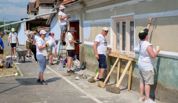 Group of volunteers painting the exterior wall of a building on a sunny day; one person on a ladder applying paint while others work nearby.