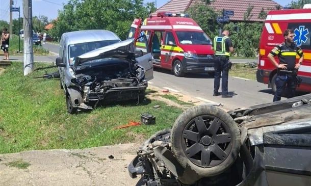 Two damaged vehicles after a crash: a silver van with severe front-end damage on the grass and a flipped car on the road, with paramedics and ambulances nearby on a sunny day.