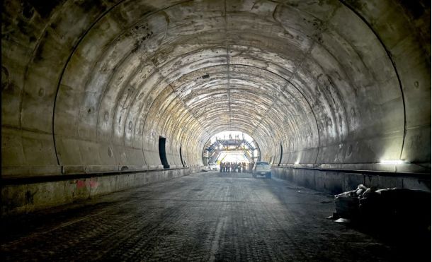Long concrete tunnel under construction; roadbed graded and workers gathered near the bright far end exit.