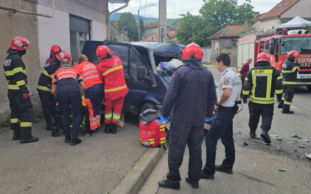 Firefighters and paramedics surround a severely damaged blue van after a street crash, assisting a patient as a crowd stands nearby with a fire truck visible in the background.