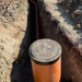 Open trench with an orange PVC pipe and a circular manhole/access lid at a construction site.